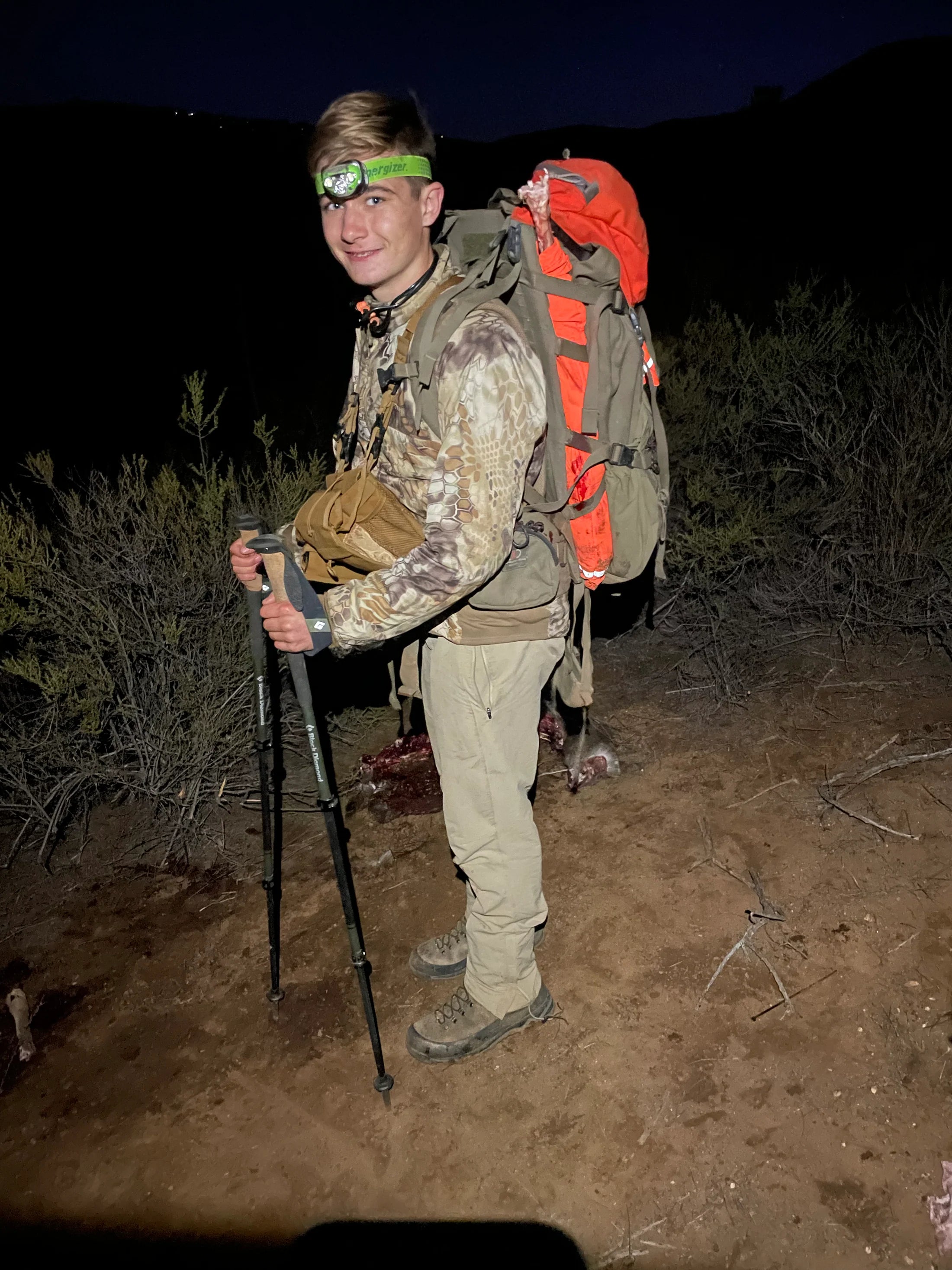 Young man in camouflage clothing with hiking gear and TRICER game bags strapped to his back at night